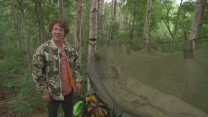 Foto van presentator in camouflagekleding bij een soort legertent, afdak van tentdoek, in het bos.