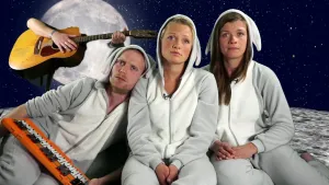 Foto van een man en twee vrouwen die naast elkaar zitten met witte kleding en capuchons op, op de achtergrond zie je iemand die gitaar speelt.