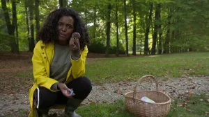 Foto van presentator die gehurkt zit op de grond in het bos en bekijkt een paddenstoel die ze in haar hand houdt.