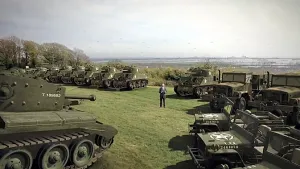 Foto van een grasveld met veel tanks erop, in het midden staat een man, presentator.