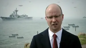 Foto van presentator met op de achtergrond grote oude foto, zwart/wit, met zee met bootjes en een oorlogsschip erop.