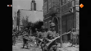 Oude foto, zwart/wit, van soldaten met geweren die door een straat lopen met oorlogsschade.