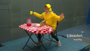 Foto van jongen in gele regenkleding die zit aan een rood/wit gedekte tafel met daarop een boek, waterkan, bordje.