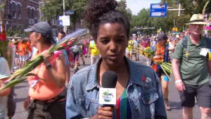 Foto van presentator met microfoon die staat op de weg waar de wandelaars van de Nijmeegse Vierdaagse aan komen, aantal houden gladiolen vast.