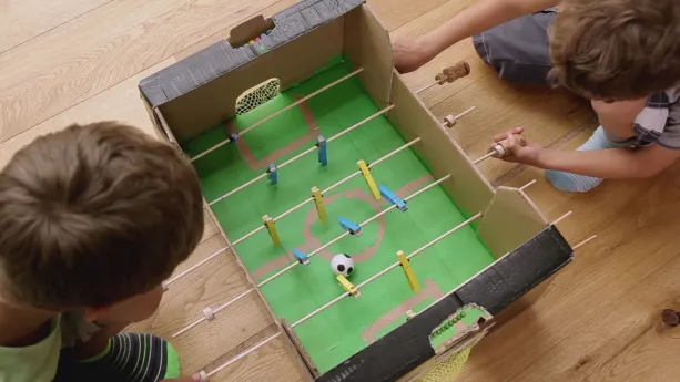 Foto van een zelfgemaakt voetbaltafelspel van met name karton waarmee twee jongetjes spelen.