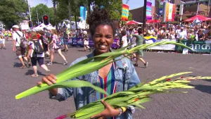 Foto van presentator met gladiolen in haar hand met op de achtergrond de finish van de Nijmeegse Vierdaagse waarbij wandelaars binnenkomen en publiek.