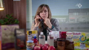 Foto van presentator die aan tafel zit met daarop allerlei zoete producten zoals appels, koekjes en limonade.
