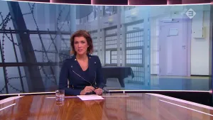 Foto van presentator die aan de desk zit met erachter foto van een TBS-kliniek van binnen, grote ruimte aldaar.
