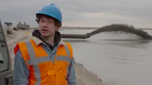 Foto van man in werkkleding langs water en zand, wadden, op de achtergrond een grote horizontale buis die slib spuit het water in.