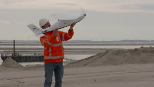 Foto van man in werkkleding die een drone oplaat bij water en zand, wadden, deze drone heeft iets weg van een vliegtuigje.