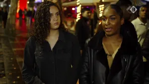 Foto van een drukke straat 's avonds, in het uitgaansleven, waar vele mensen lopen en op de voorgrond lopen twee vrouwen naast elkaar.