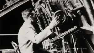 Oude foto, zwart/wit, van Edwin Hubble die zit voor een grote telescoop en erin kijkt.