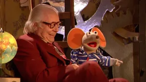 Foto van Sesamstraat met de presentator die op een stoel zit en Ieniemienie zit naasst hem, in de studio.