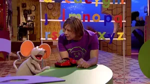 Foto van Sesamstraat waarbij presentator met Ieniemienie aan tafel zit met een ouderwetse telefoon erop.