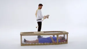 Foto van een vrouw die op haar rug ligt in soort doorzichtige doodskist, erbij staat een man met een stokpaardje tussen de benen.