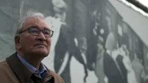 Foto, portret, van een man met bril op die staat bij een grote foto achter hem, maar niet goed te zien wat er op die foto staat.