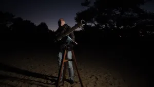 Foto van een man die 's nachts met heldere hemel buiten staat bij een kleinere telescoop die op de grond staat.