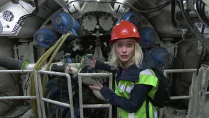Foto van presentator in werkkleding en helm op die staat bij het uiteinde van een grote machine, een tunnelboor.