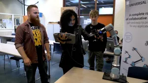Foto van man, vrouw en jongen die in een lokaal staan bij een robotarm die staat op een tafel.