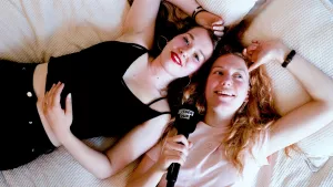 Foto van twee vrouwen die samen op de rug op een bed liggen, de ene spreekt in een microfoon.