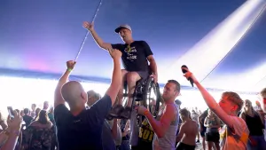 Foto van een invalide persoon in rolstoel die omhoog wordt getild door andere mensen in een tent op een festival.