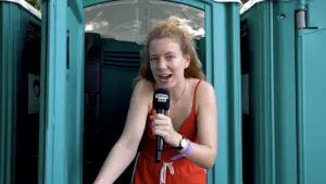 Foto van vrouw die staat bij een verplaatsbare WC, WC-hokje, die iets vertelt in microfoon.