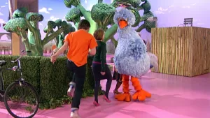 Foto van Sesamstraat met Pino in beeld, een vrouw, een jongen die huppelen, in de studio met nagemaakte bomen etc.