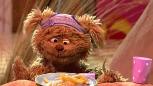 Foto van Sesamstraat waarbij Tommie aan de desk zit met een bordje met friet voor hem.