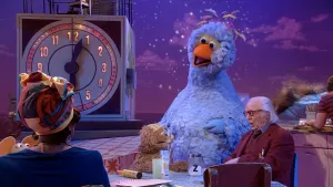 Foto van scène uit Sesamstraat waarbij pesentator en iemand anders aan tafel zitten, ook Pino en paar andere figuren.