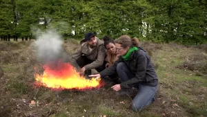 Foto van aantal mensen die gehurkt zitten op grasveldje in het bos bij een brandend vuurtje.