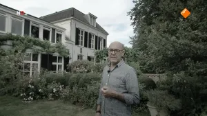 Foto van NOS-Journaal waarbij presentator/nieuwslezer staat voor een wit gebouw/groot huis met tuin.