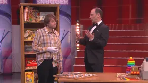 Foto van scène uit Studio Snugger met presentator en een jongen die staan in de studio bij tafel met spelletjes erop.