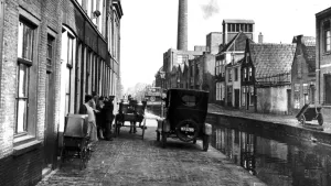 Oude foto, zwart/wit, van een straat met woningen en fabrieken, mensen en auto's, in de crisisjaren dertig