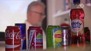 Foto van rij blikjes met frisdrank en een frisdrankfles, vaag op de achtergrond zie je een man met bril op.