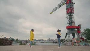 Foto van twee personen die buiten op de kade staan bij water en een hijskraan.