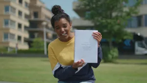 Foto van presentator op grasveld bij flatgebouw die een script, boek, vasthoudt.
