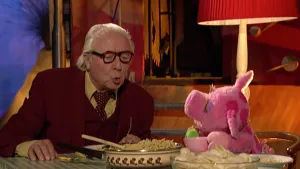 Foto van scène uit Sesamstraat met de presentator en figuur (roze biggetje, pop) aan tafel met eten erop.