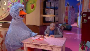 Foto van scène uit Sesamstraat waarop Pino zit aan tafeltje waaraan ook een man zit, in speelkamer.