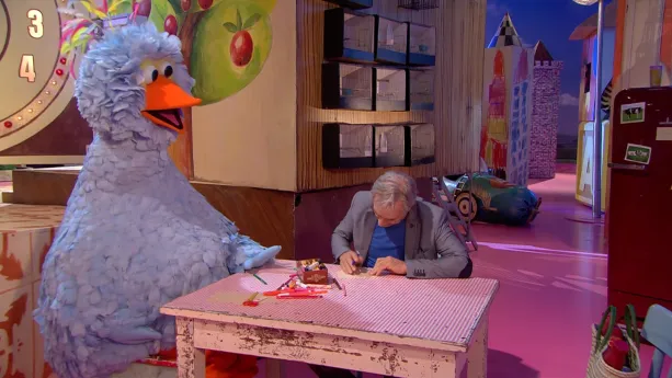 Foto van scène uit Sesamstraat waarop Pino zit aan tafeltje waaraan ook een man zit, in speelkamer.