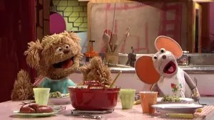 Foto van Sesamstraat met Tommie en Ieniemienie (poppen) zittend aan tafel, op tafel staan borden met boerenkool, een pan etc.