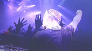 Foto van een discotheek met gekleurde verlichting, je ziet de Dj van achteren en omhoog gestoken handen van de dansende mensen.