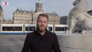 Foto van NOS-Journaal met presentator, op de achtergrond de Dam te Amsterdam, Paleis op de Dam, tram die er voorbij rijdt.