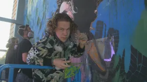 Foto van presentator, achter hem twee personen die bezig zijn aan een grote muurschildering.