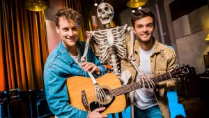 Foto van twee mannen in studio, één van hen speelt op een gitaar, tussen hen in staat een skelet/geraamte.