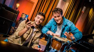Foto van twee mannen in kamer met muziekinstrumenten, de ene heeft soort instrumentje in zijn hand, de andere trommelt op een trommel (djembe).