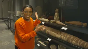 Foto van presentator die staat in museum bij kast met allerlei beelden, stenen, mummies etc. erin.