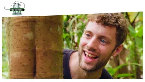 Foto van lachende man, je ziet zijn hoofd, die staat bij een boom waarvan je de stam ziet.