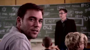 Foto van man, je ziet zijn hoofd, in een klas, je ziet nog paar kinderen, de leraar die staat voor het schoolbord.