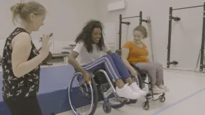 Foto van vrouw in een rolstoel die daarmee oefeningen/kunstjes doet, ernaast andere vrouw in rolstoel, erbij staat een vrouw die toekijkt, in een sportzaal.
