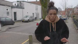 Foto van presentator in straat met huizen die iets aan het vertellen is.